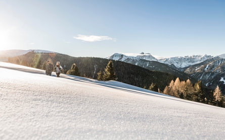 A man and a woman making a winter hike in the snow, in the background the Peitlerkofel peak and other snow covered mountains