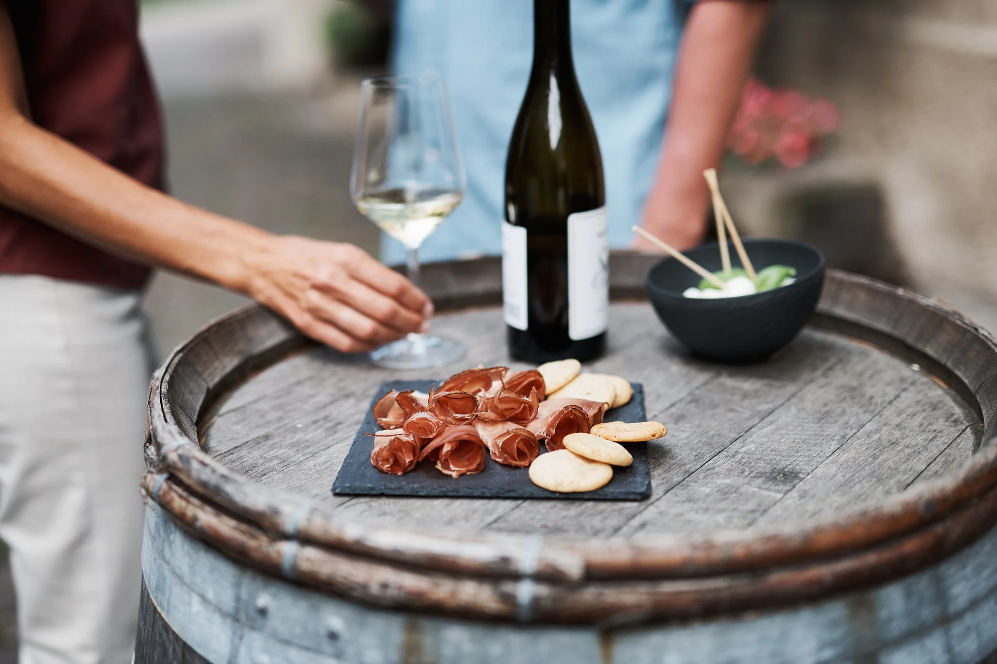 Zwei Personen stehen neben einem Fass auf dem ein Glas Weißwein, eine Flasche Weißwein und eine Platte mit Speck stehen