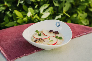 Ein Teller mit cremigem Getreidegericht, belegt mit dünnen Apfelscheiben, frischen Kräutern und violetten Blättern.