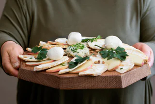 Ein rustikales Holzbrett mit Mozzarellakugeln, Apfelscheiben, Käse, Pinienkernen und frischer Petersilie.