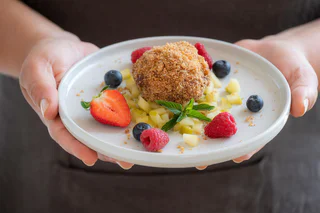 Ein mit Bröseln bedeckter Knödel auf Apfelwürfeln, umgeben von Beeren und Minzblättern.