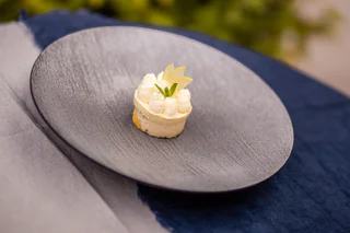 Kleines, rundes Dessert mit heller Creme, dekoriert mit Apfelscheiben und Minzblatt auf dunklem Teller.