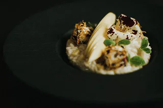 Cremiges Risotto mit Apfelscheiben, Kräutern, Blüten und knusprigen Häppchen auf dunklem Teller.