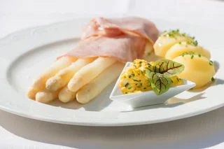 Weißer Spargel mit gekochtem Schinken, kleinen Kartoffeln mit Schnittlauch und einer Portion Bozner Sauce, angerichtet auf einem weißen Teller.