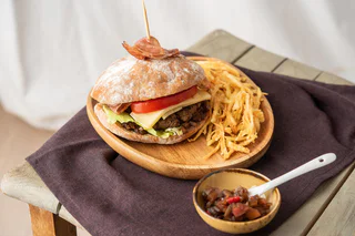 Rustikaler Burger mit Salat, Tomate, Käse und Speck im Brötchen, serviert mit knusprigen Kartoffelsticks und einer kleinen Schale Chutney.
