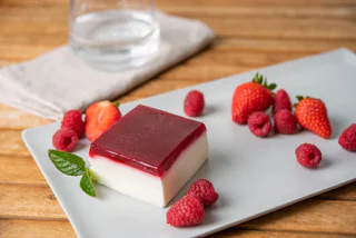 Quadratisches Schichtdessert mit rotem Beerenspiegel auf weißer Panna-Cotta, serviert mit frischen Erdbeeren, Himbeeren und Minze auf einem Teller.