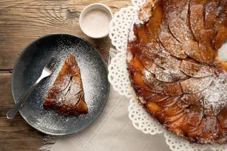 Karamellisierter Apfelkuchen mit Puderzucker bestreut, ein Stück auf einem dunklen Teller mit Gabel, daneben eine kleine Schale mit Sauce.
