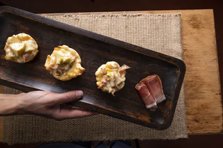 Drei kleine herzhafte Törtchen mit geschmolzenem Käse, Speck und Gemüse auf einem dunklen Holzbrett, daneben zwei aufgerollte Speckscheiben.