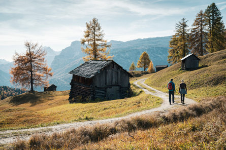 Ein Mann und eine Frau spazieren zwischen Holzhütten über einen Weg in der Region Alta Badia.