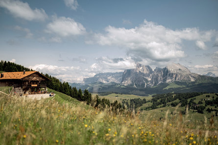 Vista panoramica da una malga di montagna sull'Alpe di Siusi. Diverse persone siedono a mangiare ai tavoli esterni della malga.