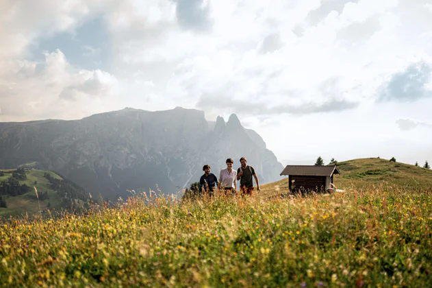 Drei Freunde wandern über die blühenden Wiesen der Seiser Alm, hinter ihnen eine Hütte und im Hintergrund das Schlernmassiv