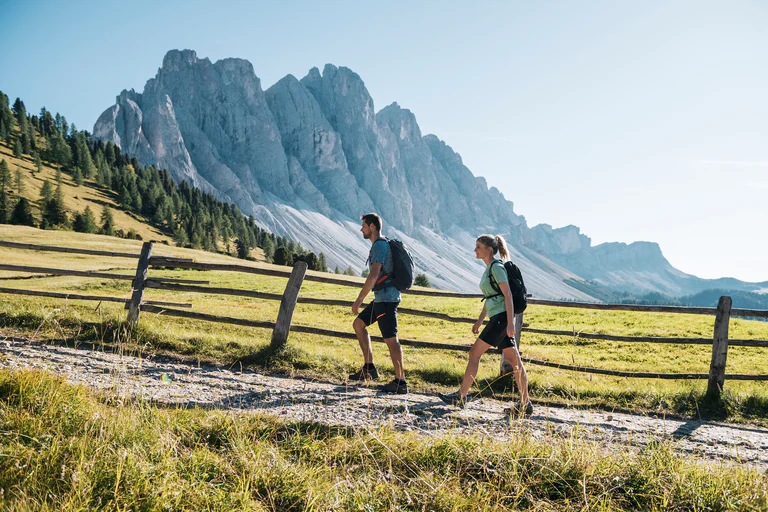 Due persone camminano su un sentiero di montagna con alle spalle le Odle in Val di Funes