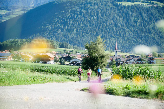 Drei Radfahrer von hinten gesehen radeln auf einem asfaltierten Teilstück des Radweges dem Dorf Olang entgegen.