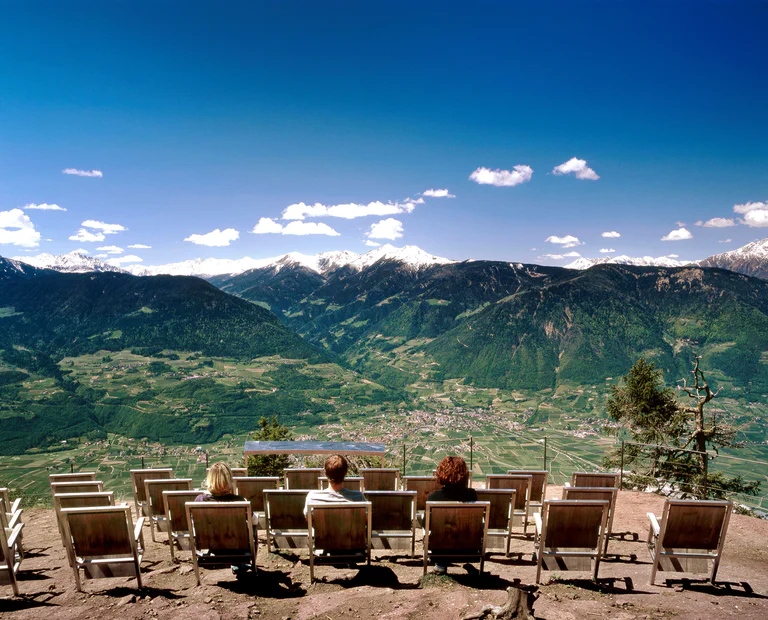 Deux femmes et un homme admirent le panorama sur la ville de Meran depuis un point de vue avec de nombreuses chaises en bois