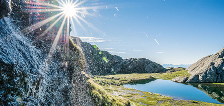 Uitzicht op een bergmeertje omgeven door rotsen. De zon schijnt en tovert lichtreflecties.