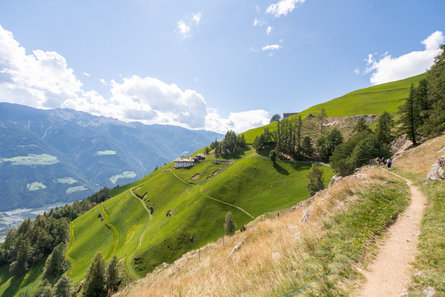 Un uomo e una donna camminano uno dietro l'altro in un prato verde. Sullo sfondo si intravedono montagne verdi e il paese di Naturno.