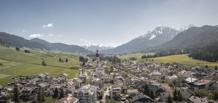View of Niederdorf/Villabassa surrounded by green meadows, fields and partly snow covered mountains.