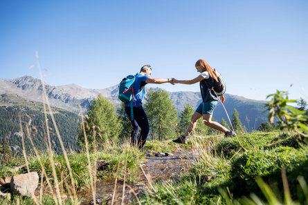 Una coppia di escursionisti cammina circondati dai paesaggi montani della Val d'Ultimo, l'uomo aiuta la donna ad attraversare un corso d'acqua tendendole la mano