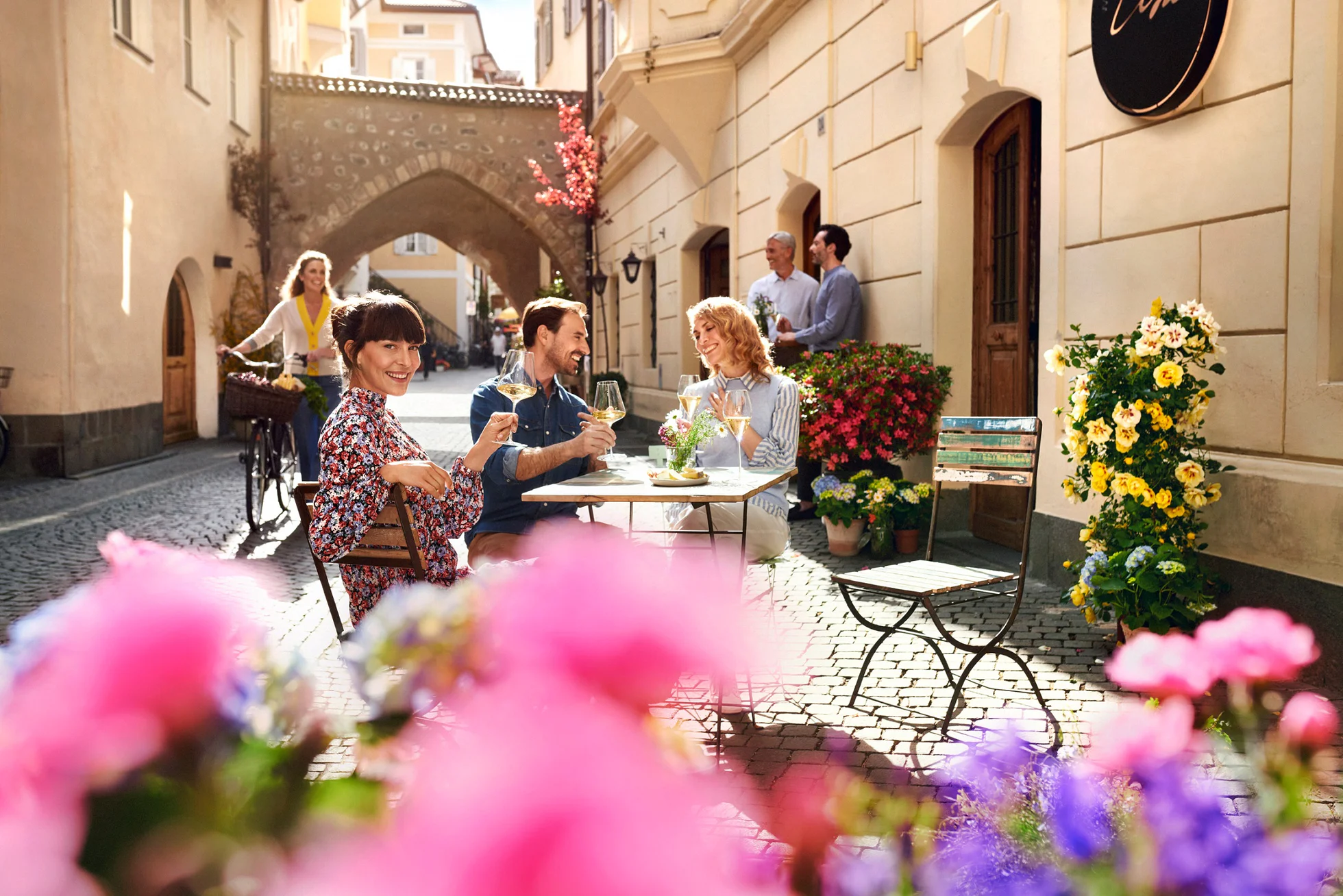 Tre persone fanno un aperitivo con calici di vino bianco nel centro storico di Bolzano decorato da una moltitudine di fiori colorati