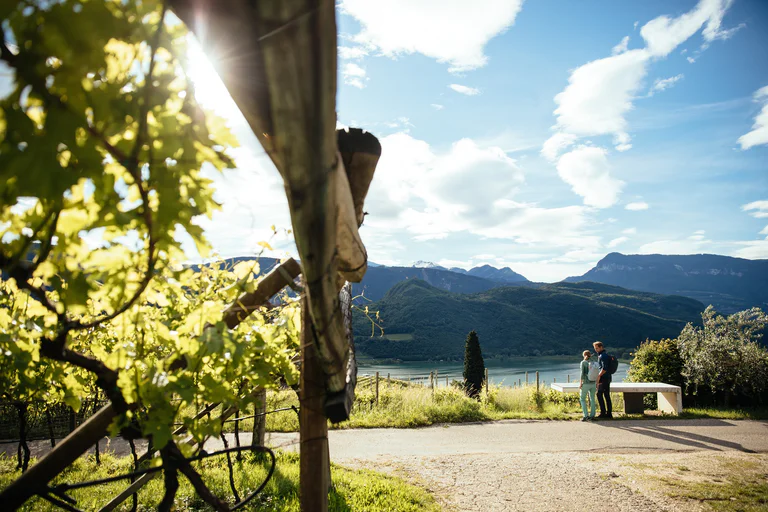 Una coppia in tenuta da trekking davanti al Lago di carezza con un paesaggio di vigneti al sole