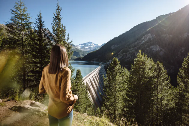 Une femme brune aux cheveux mi-longs admire un barrage dans un cadre de montagnes et de forêts de conifères.