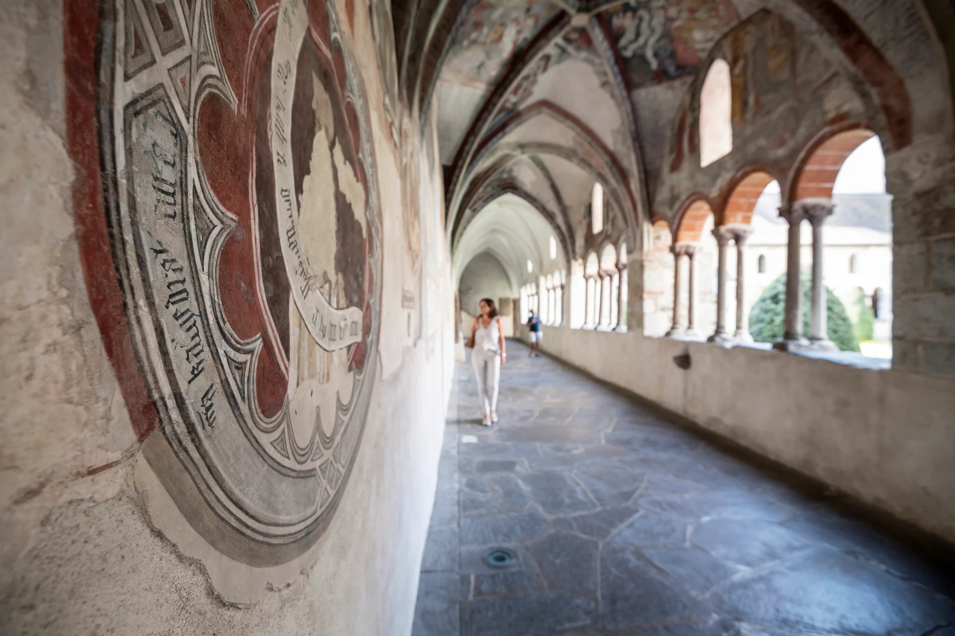 Frescoes from the 14th century adorn the cloister of Brixen Cathedral. A visitor is in the background.