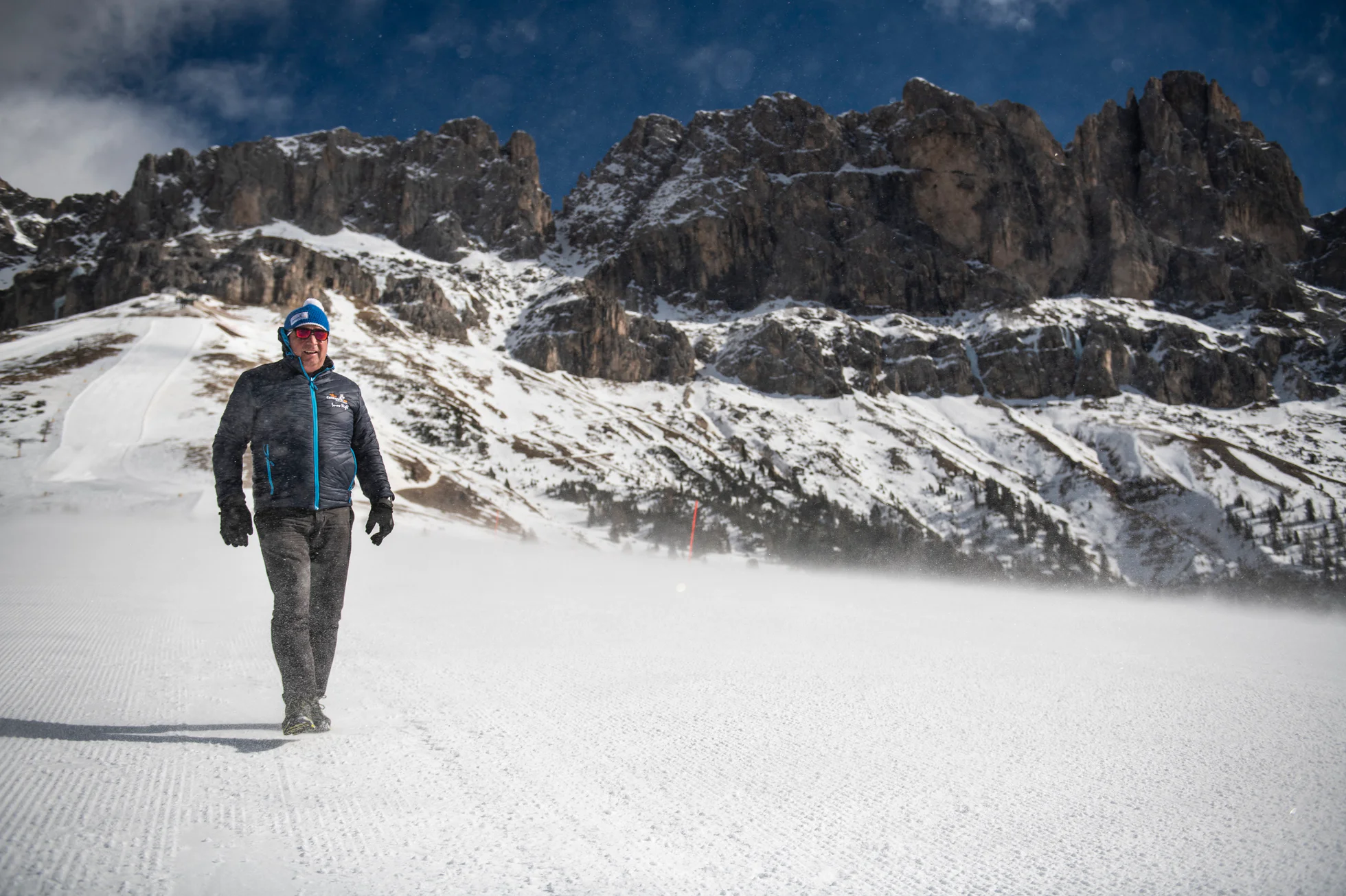 Snow expert Georg Eisath strides across an expanse of snow in front of a mountain panorama.