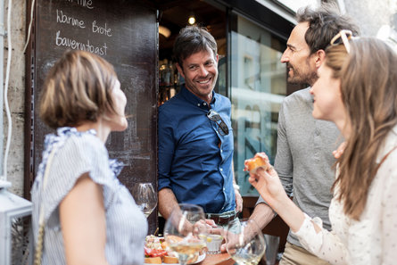 Ein typischer Genussmoment in einer Bar im Herzen von Bozen: Zwei Frauen und zwei Männer entspannen bei einem Aperitivo sowie klassische Südtiroler Spezialitäten wie Speck.