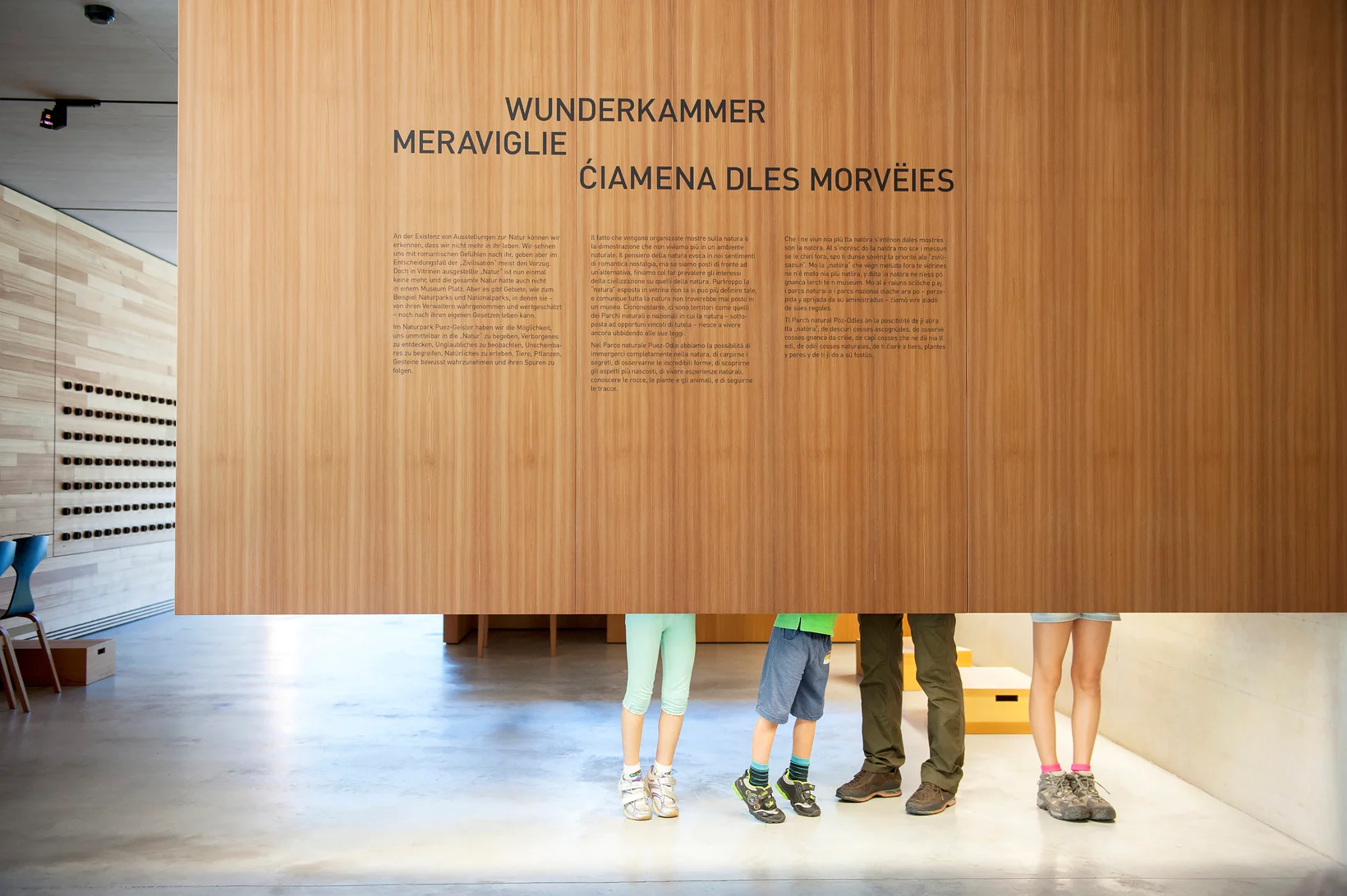 Le gambe di quattro visitatori della Casa del Parco Naturale di Funes sono dietro una parete espositiva con le tre lingue altoatesine: italiano, tedesco e ladino.