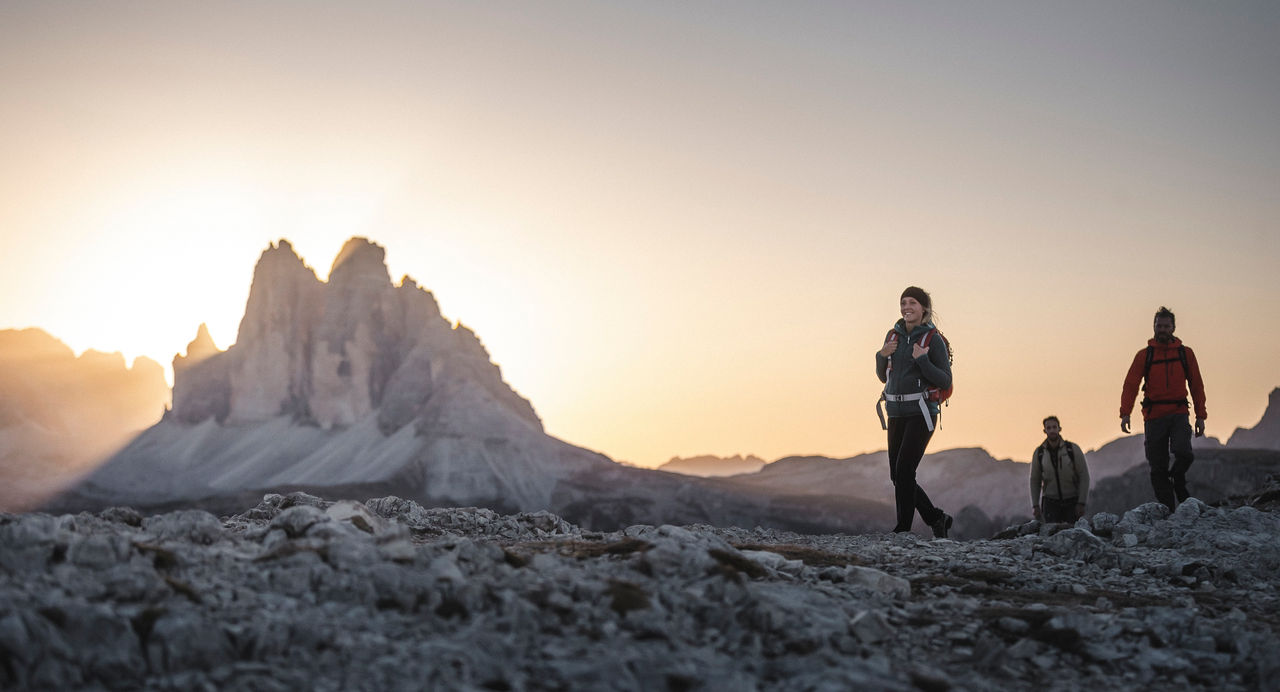 Zwei Manner und eine Frau wandern im Hochgebirge, während die Sonne hinter den Dolomiten versinkt