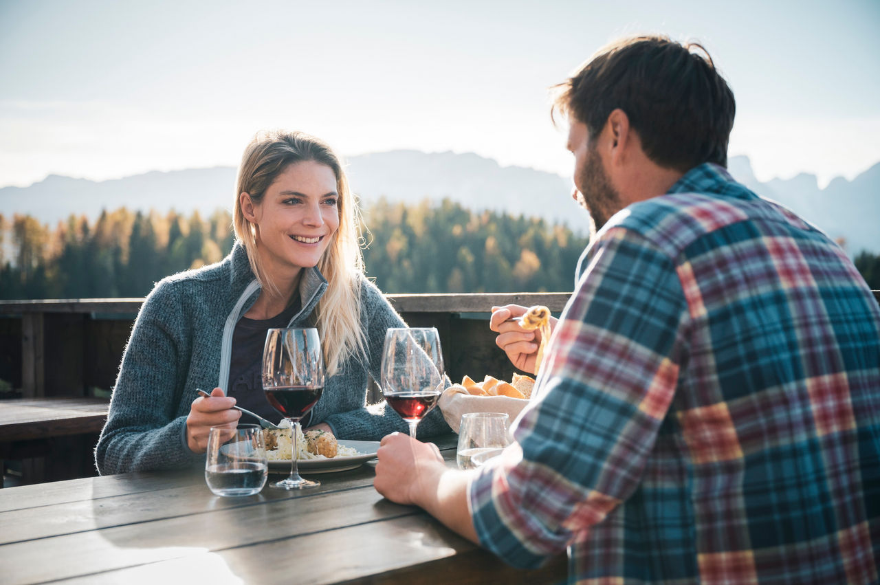 Eine Frau lächelt und sitzt mit einem Mann beim Essen auf der Hüttenterrasse in der Herbstsonne.