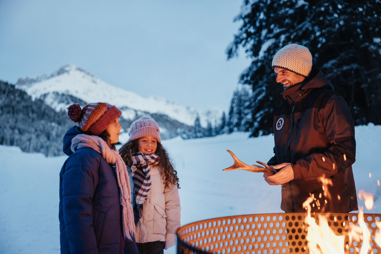 Zwei Kinder und ein Erwachsener stehen in den Abendstunden an einem Feuer im Winter