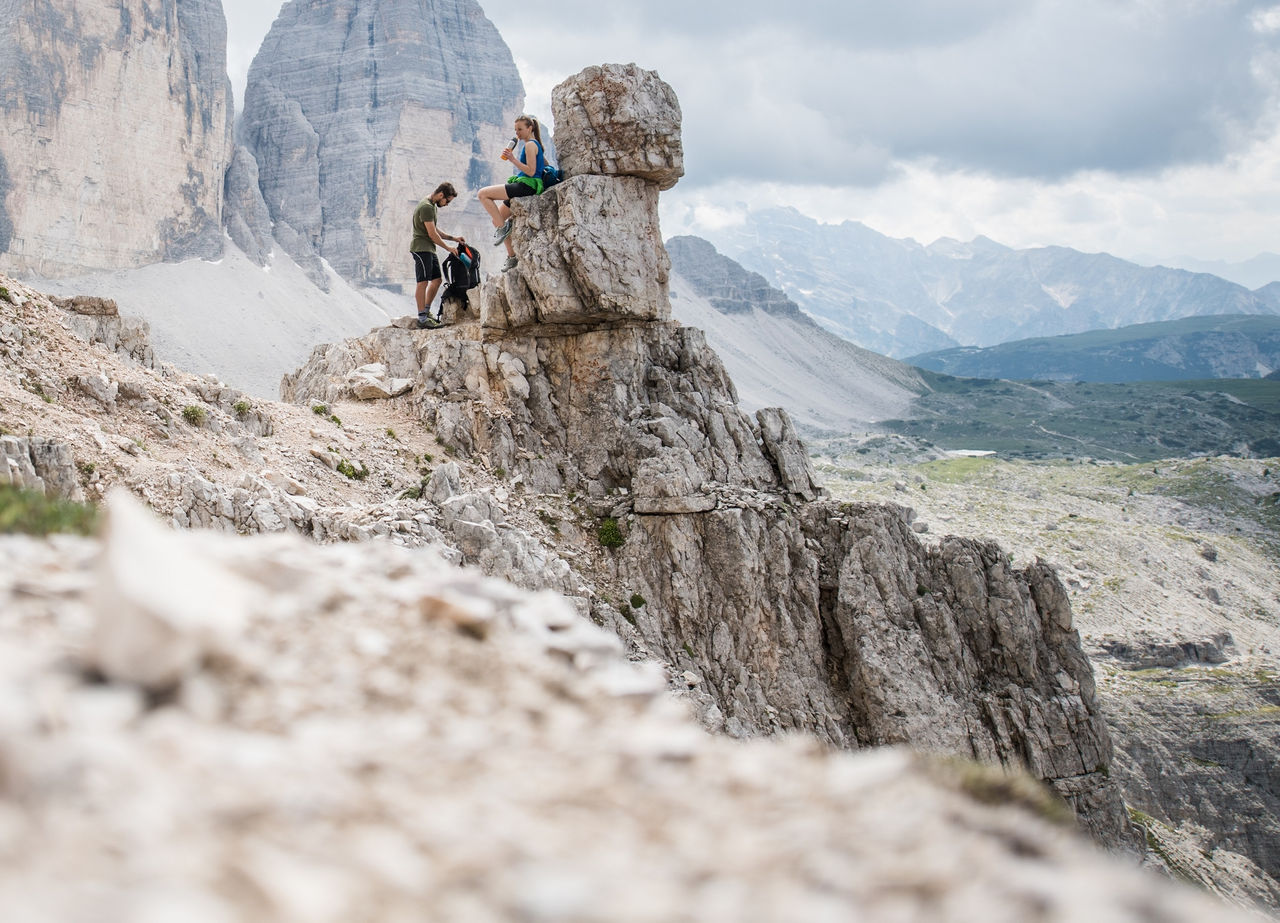 Ein Mann und eine Frau im Wanderoutfit bei einer Rast an einem Felsen im Hochgebirge mit Blick auf die Drei Zinnen
