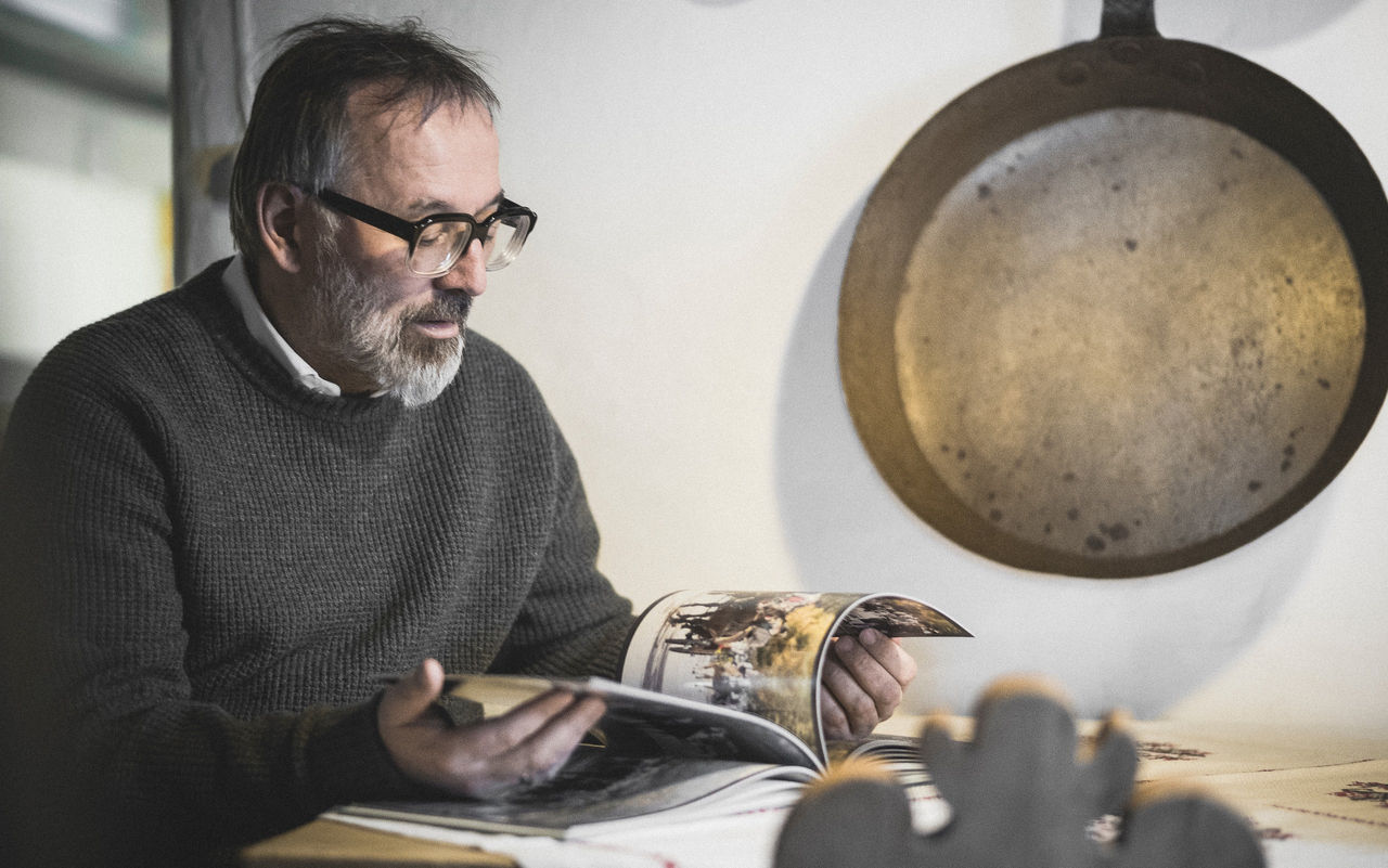 Sternekoch Norbert Niederkofler sitzt an einem Tisch und blättert in einem Bildband. Neben ihm hängt eine große Pfanne.