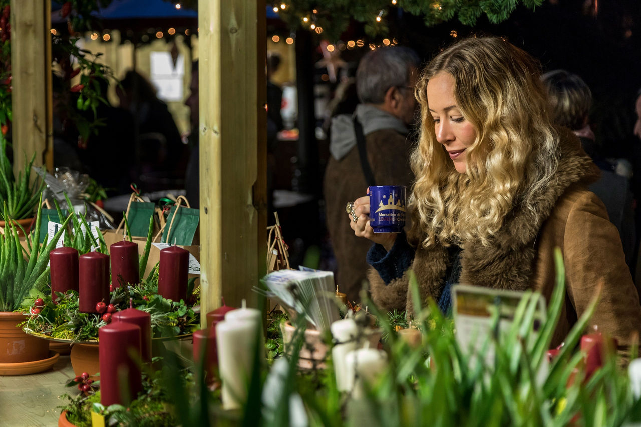 Frau mit einer Tasse in der Hand steht vor einem Verkaufsstand mit Adventskränzen