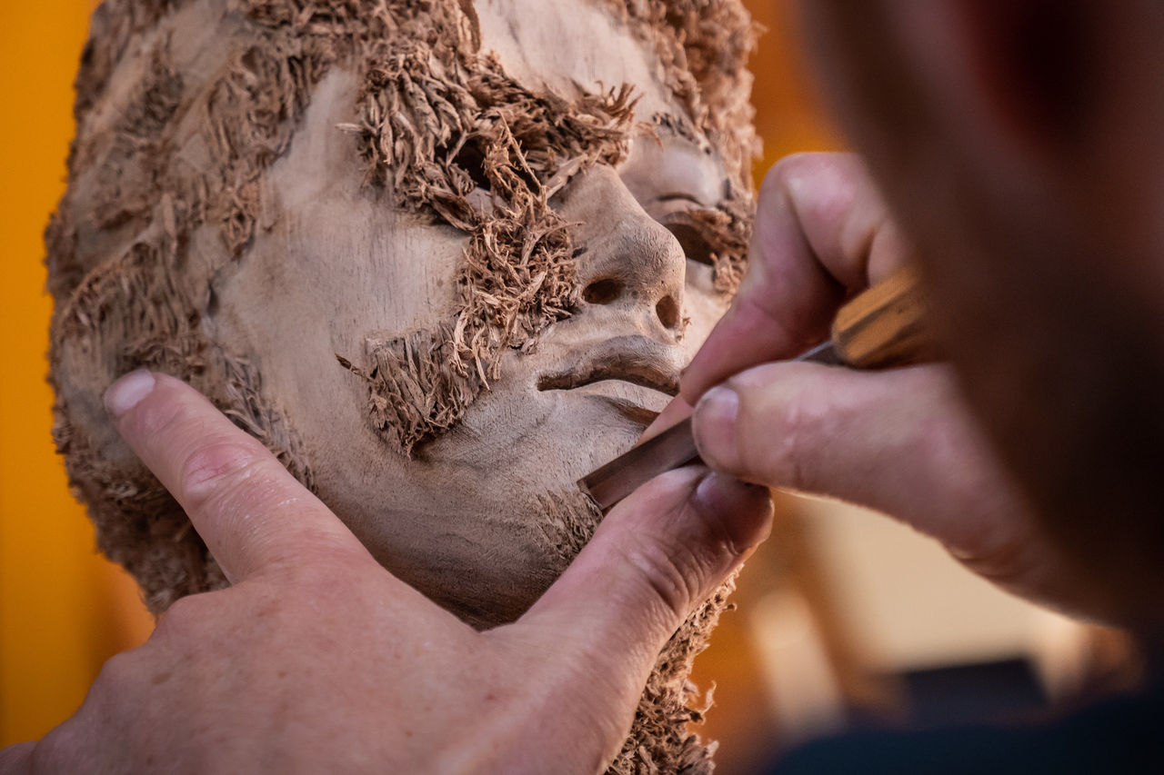 Die Hände von Bildhauer Aron Demetz bearbeiten mit einem sogenannten Pfeil das Kinn einer Holzskulptur, die gerade entsteht.