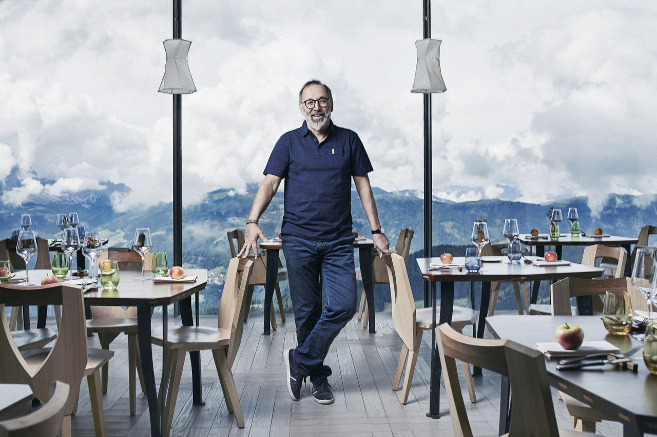 Der Koch Norbert Niederkofler steht zwischen den Tischen seines Restaurants AlpiNN - hinter ihm geht der Blick durchs Fenster auf die Berge.