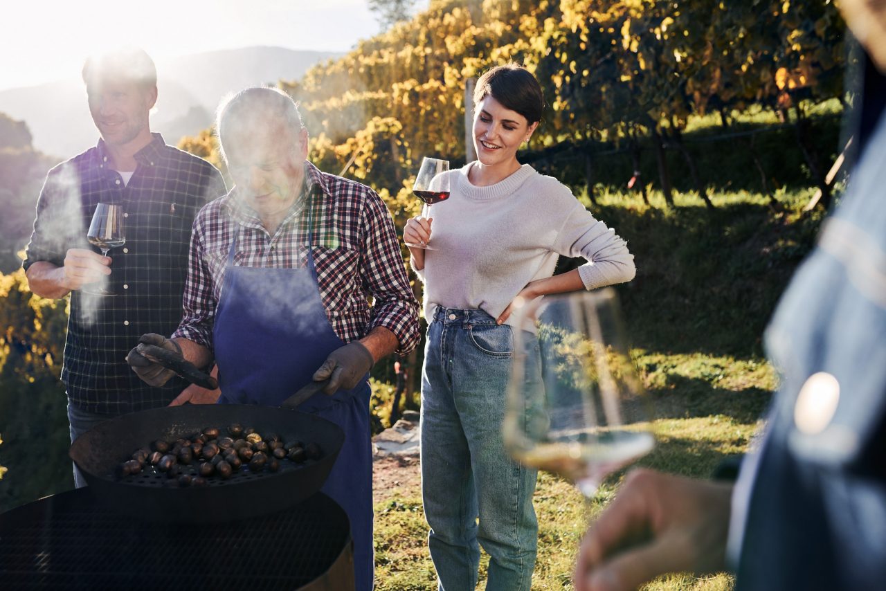 Beim Törggelen an einem sonnigen Herbsttag im Eisacktal steht eine Gruppe von Menschen mit Weingläsern in den Händen und betrachtet das tradtionelle Kastanien-Rösten. 