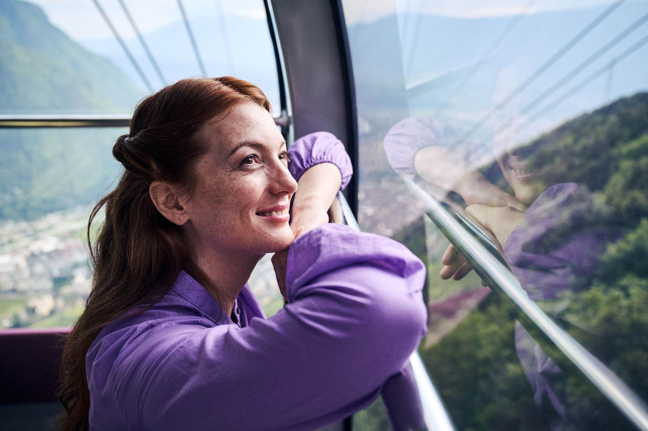 Eine junge Frau lächelt und blickt aus der Gondel der Rittner Seilbahn, die Stadt Bozen im Hintergrund