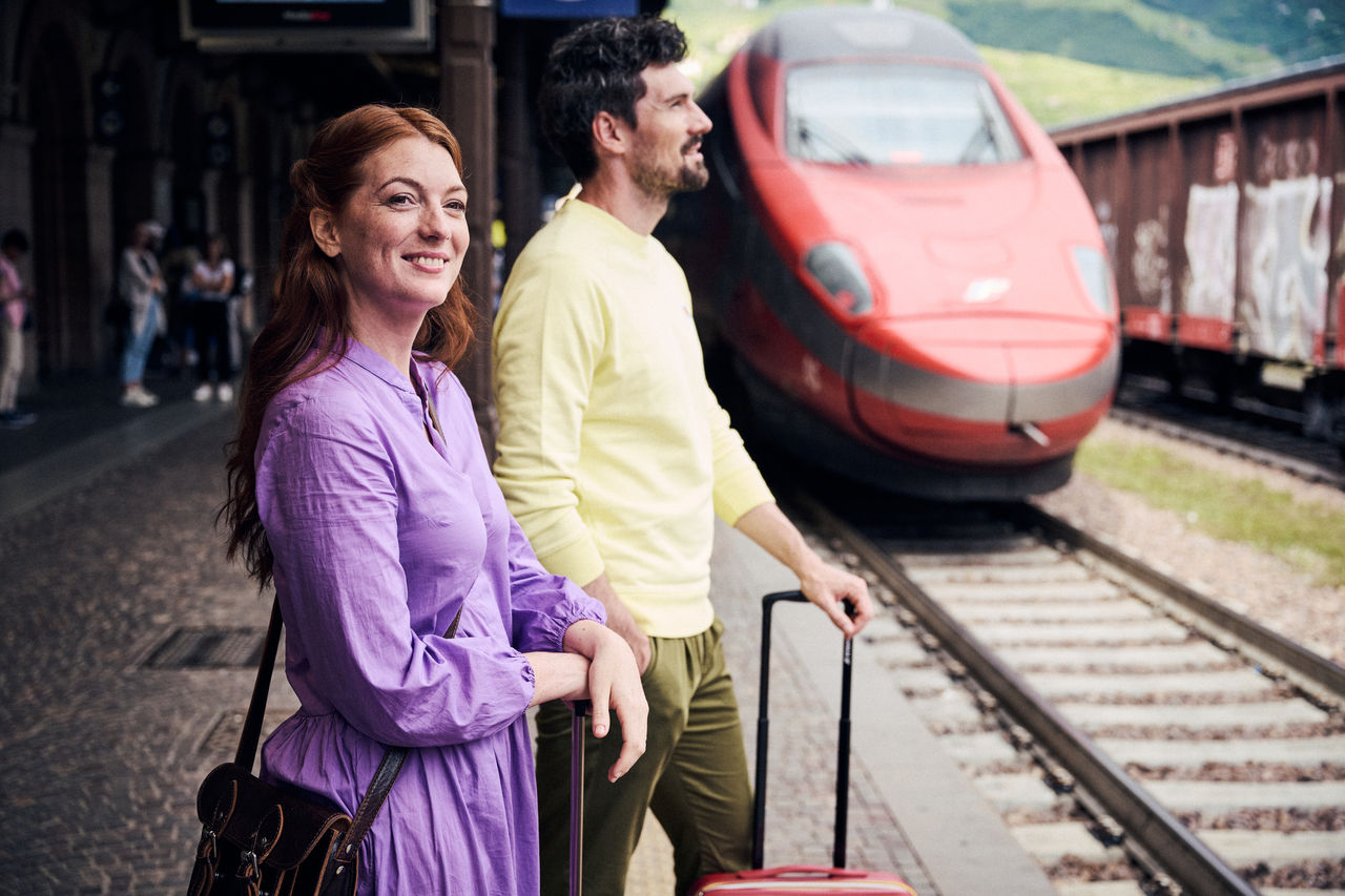 Ein junges Paar steht lächelnd am Bahngleis, während ein Schnellzug einfährt.