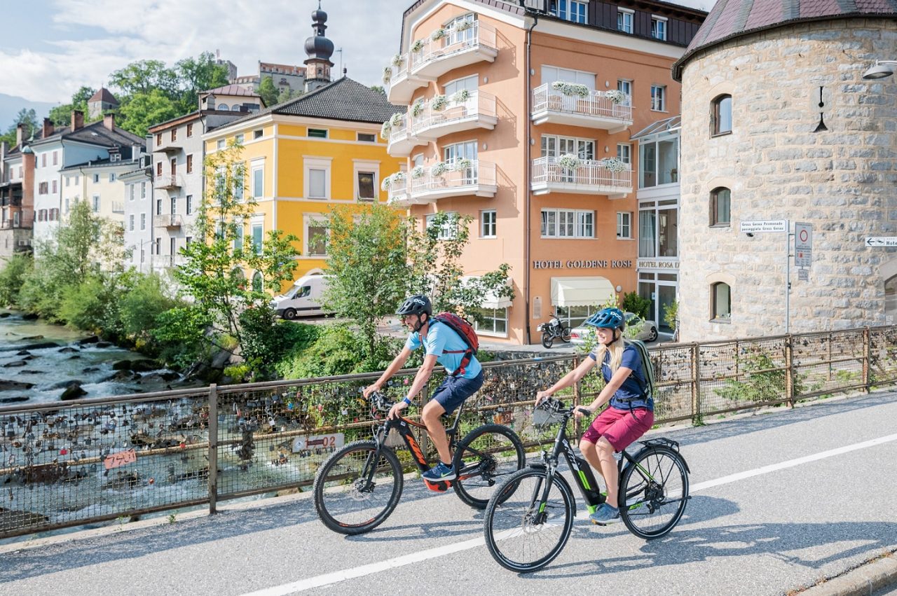 Ein Mann und eine Frau auf dem Rad fahren über eine Brückein Bruneck, im Hintergrund die bunten Häuserfassaden der Altstadt