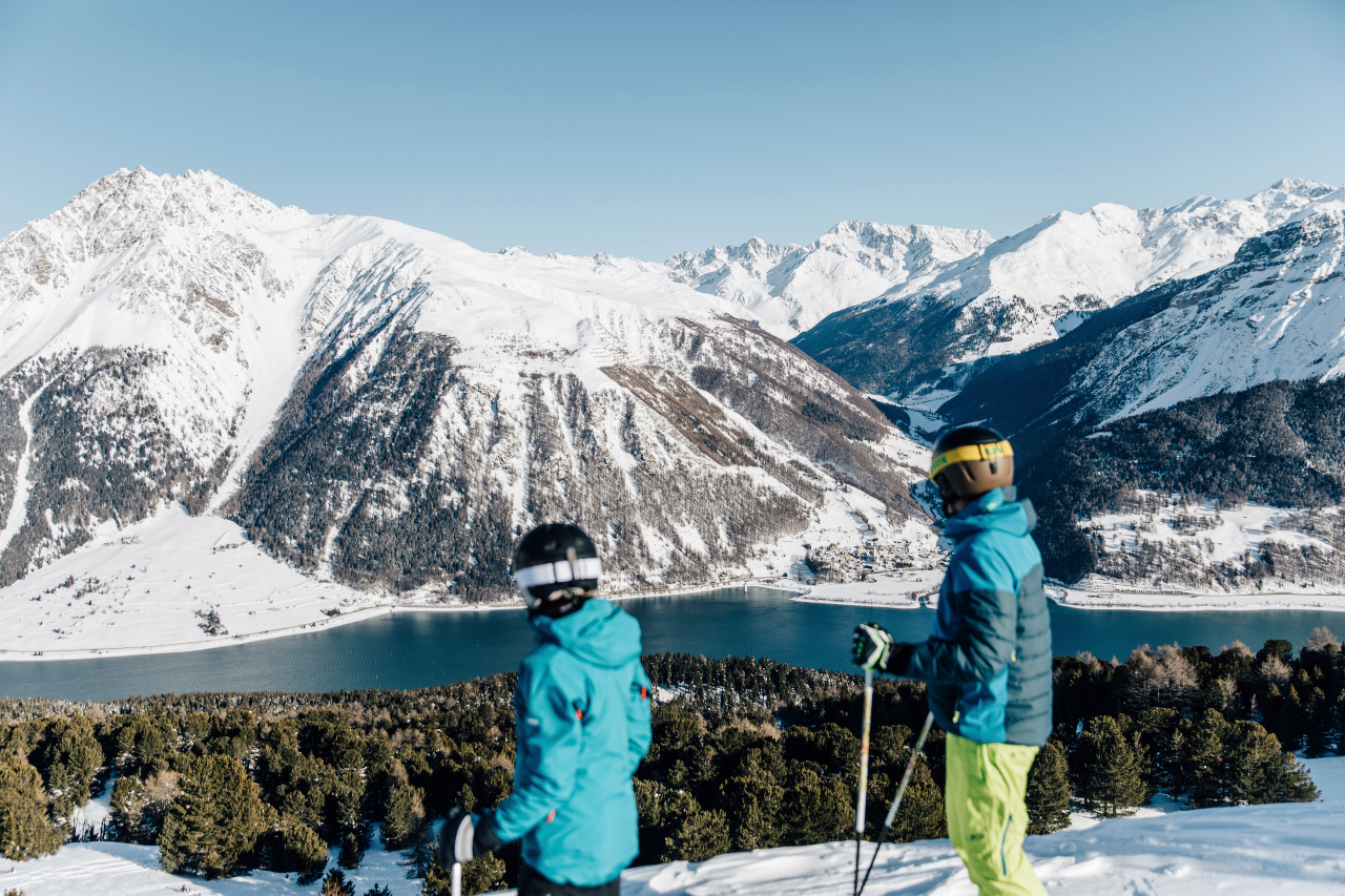 Zwei Skifahrer blicken von der Haider Alm auf den Reschensee, der von schneebedeckten Gipfeln eingerahmt ist.