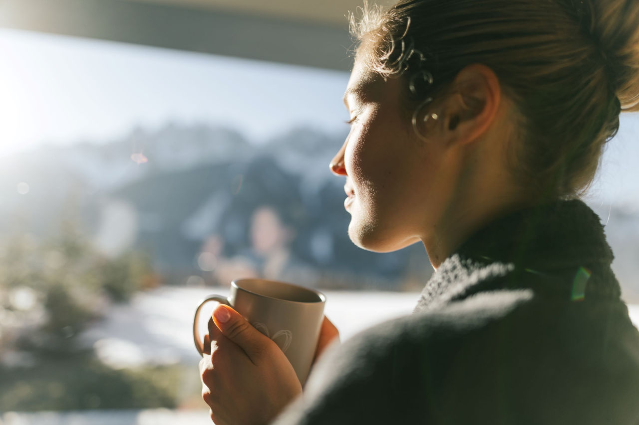 Eine junge Frau steht entspannt am Fenster mit einer Teetasse in der Hand.