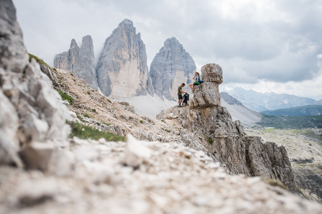 Ein Mann und eine Frau im Wanderoutfit bei einer Rast an einem Felsen im Hochgebirge mit Blick auf die Drei Zinnen