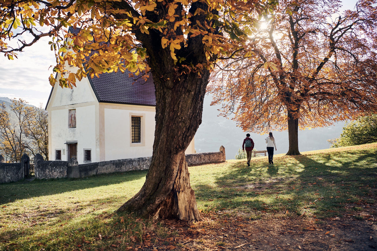 Ein Mann und eine Frau wandern im Herbstlicht über eine Wiese neben einer Kapelle.