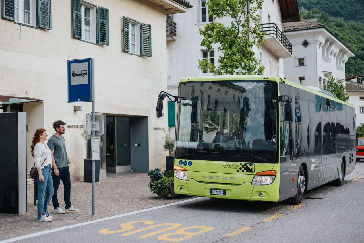 Ein junges Paar wartet an der Bushaltestelle in einer charmanten Straße, während ein Bus der regionalen Linien einfährt.