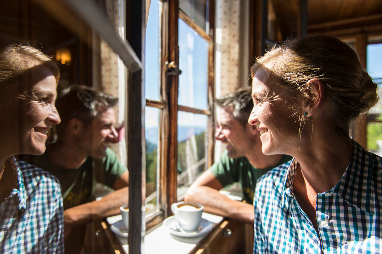 Eine Frau und ein Mann schauen bei Sonnenschein aus dem Fenster. Zwischen ihnen steht eine Tasse Cappuccino und am linken Bildrand spiegelt sich das Gesicht der Frau in einer Scheibe.