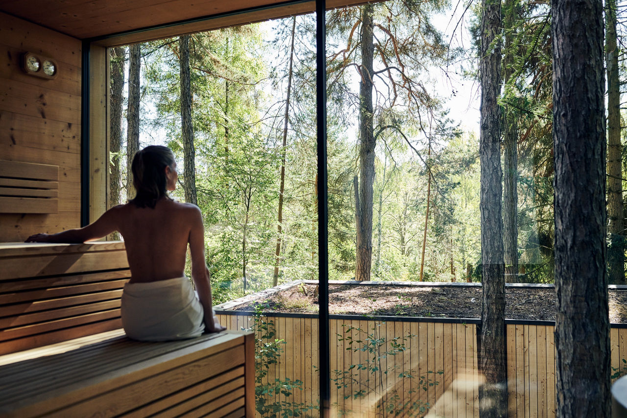 Eine Person sitzt in der Sauna und blickt durch ein Panorama Fenster auf den Wald im Hintergrund